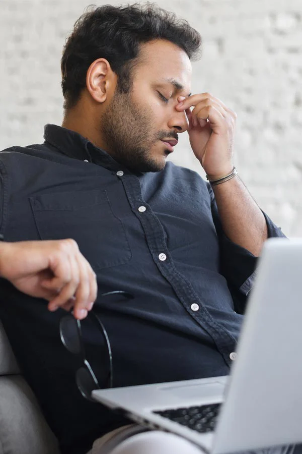 A middle-aged man with a laptop closing his eyes and rubbing his nose. He needs treatment for Adrenal Fatigue Syndrome from Tanya Zboril, NP of TBT Medical in Edmonton.