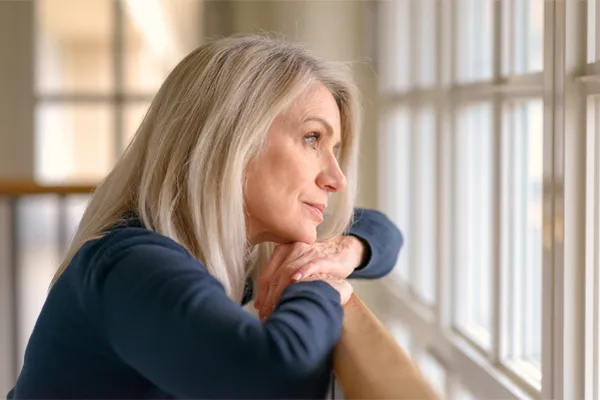 A mature woman in a blue shirt looks out the window with melancholy. Schedule treatment for adrenal hormone disorder from Tanya Zboril, NP of TBT Medical in Edmonton.
