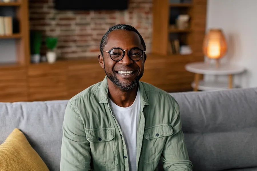 A middle aged black man in a green button up shirt sits on his couch smiling, happy with his treatment for andropause and low testosterone from Tanya Zboril, NP of TBT Medical in Edmonton.