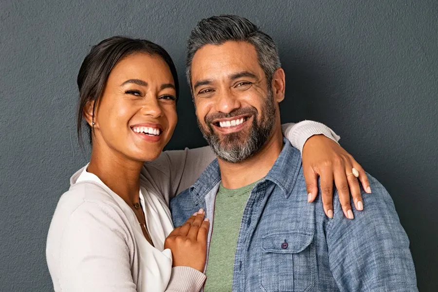 A middle-aged couple with their arms around each other standing against a gray background and smiling, happy with the bioidentical hormone therapy they received from Tanya Zboril, NP of TBT Medical in Edmonton.