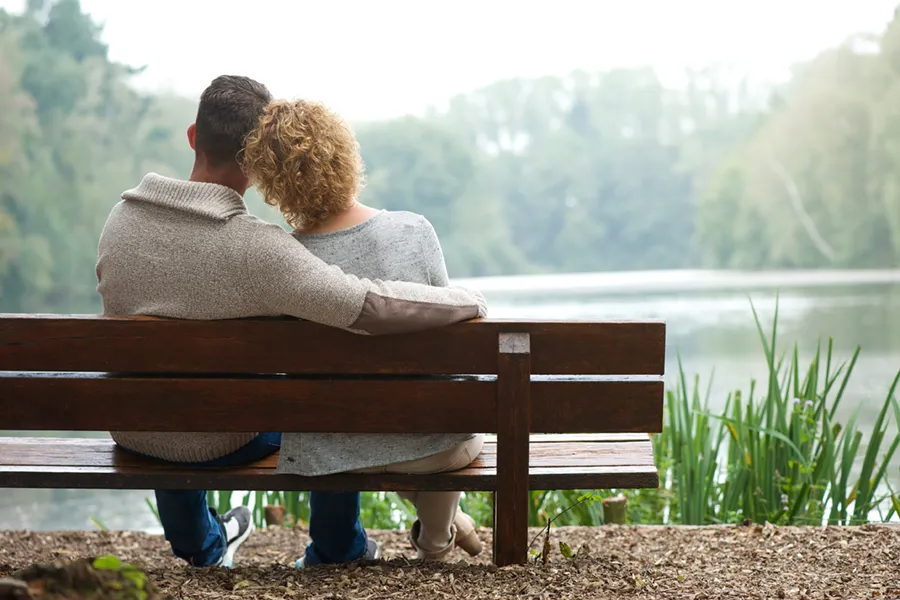 A couple wearing sweaters seen from behind sitting on a bench and looking out at a relaxing lake and forest, representing functional medicine treatment from Tanya Zboril, NP of TBT Medical in Edmonton.