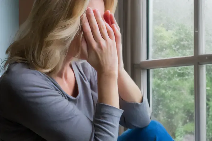 A blonde woman in a gray shirt sits in front of a window on a rainy day covering her eyes with her hands. Get treatment for Grave's Disease from Tanya Zboril, NP of TBT Medical in Edmonton.