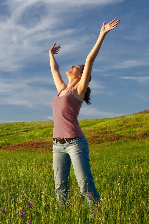 A woman in a tank top and jeans looks up with her arms outstretched to the sky in a field after successful treatment for Grave's Disease from Tanya Zboril, NP of TBT Medical in Edmonton.