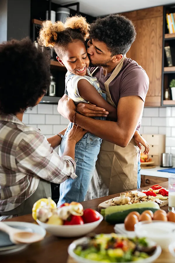Gut-Health-Treatment A man holds his daughter and kisses her cheek as the family makes a healthy dinner in the kitchen. Get help for digestive issues and gut health from Tanya Zboril, NP of TBT Medical in Edmonton.