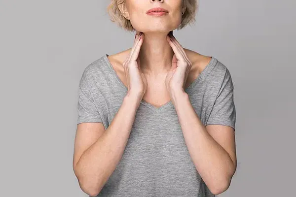 An older blonde woman in a gray t-shirt holds her hands to her neck checking for signs of thyroid problems. Get help for Hashimoto's Thyroiditis from Tanya Zboril, NP of TBT Medical in Edmonton.