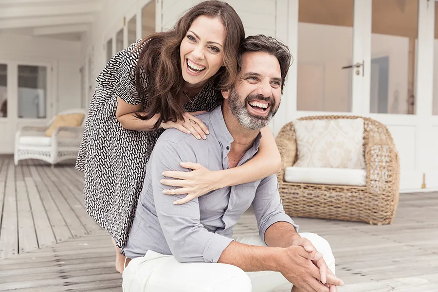 Health-and-Wellness-Services-Doctor A middle-aged couple on a wooden porch with wicker furniture; the woman is playfully hugging the man from behind. They are benefiting from the health and wellness services from Tanya Zboril, NP of TBT Medical in Edmonton.