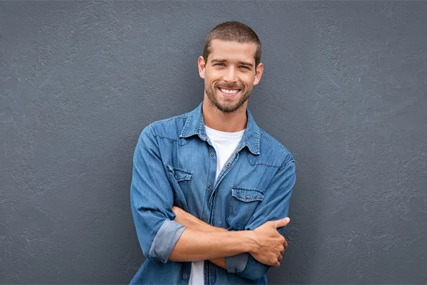 Hormones-for-Men-Doctor A man in a denim shirt stands smiling against a gray-blue wall, pleased with his testosterone hormone therapy from Tanya Zboril, NP of TBT Medical in Edmonton.
