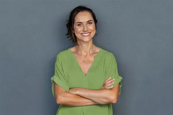 Hormones-for-Women-Doctor A woman in a green shirt stands smiling against a gray-blue wall, pleased with her hormone therapy from Tanya Zboril, NP of TBT Medical in Edmonton.