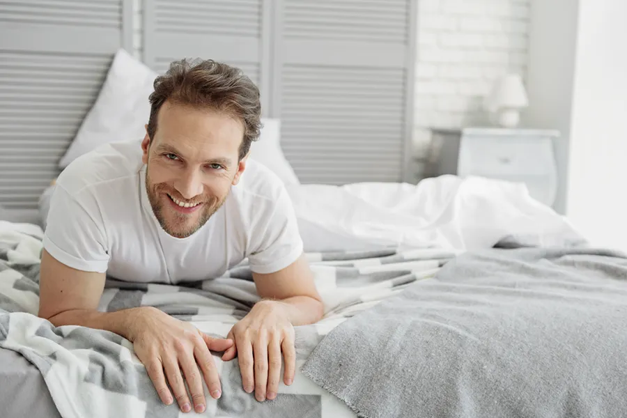 A dirty-blonde haired man lays on his stomach in bed smiling. Schedule treatment for low libido in men from Tanya Zboril, NP of TBT Medical in Edmonton.