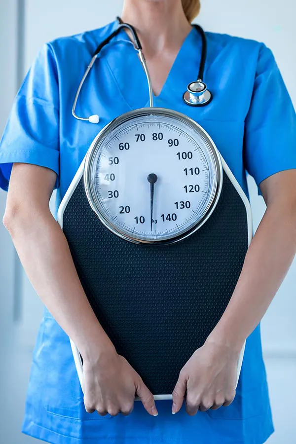 Medical-Weight-Loss-Treatment A nurse in blue scrubs with a stethoscope hanging around her neck holds a scale, representing weight loss treatments from Tanya Zboril, NP of TBT Medical in Edmonton.