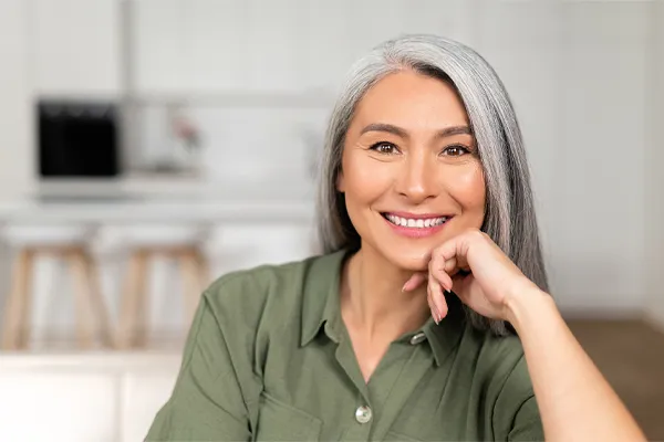 An asian woman with long gray hair and a green shirt sitting and smiling on her couch, happy with her menopause care from [NAME} in Edmonton.