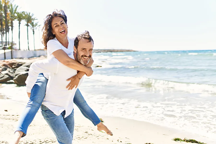 NAD-Therapy-Clinic A woman piggybacks on her husband as the middle-aged couple have fun at the beach. Get NAD Therapy from Tanya Zboril, NP of TBT Medical in Edmonton.