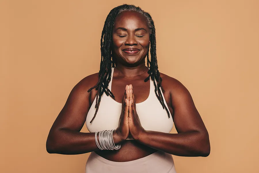 An older African American woman confidently posing against a warm background smiling with hands in a prayer pose. Tanya Zboril, NP of TBT Medical treats obesity using functional medicine in Edmonton.