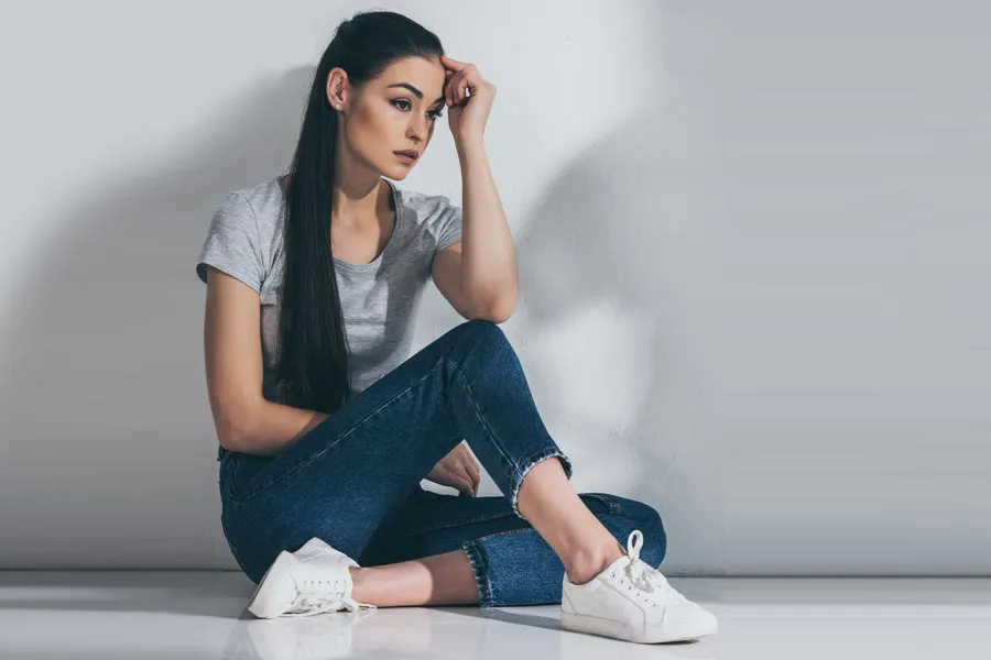 A young brunette woman in a gray t-shirt and jeans sits against a gray wall with her head resting on her hand and a distressed expression. Get help for Polycystic Ovarian Syndrome (PCOS) from Tanya Zboril, NP of TBT Medical in Edmonton.