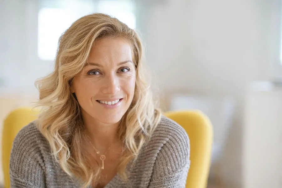 A blonde woman wearing a a gray sweater sits inside in a yellow chair, smiling and happy with her perimenopause treatment from Tanya Zboril, NP of TBT Medical in Edmonton.