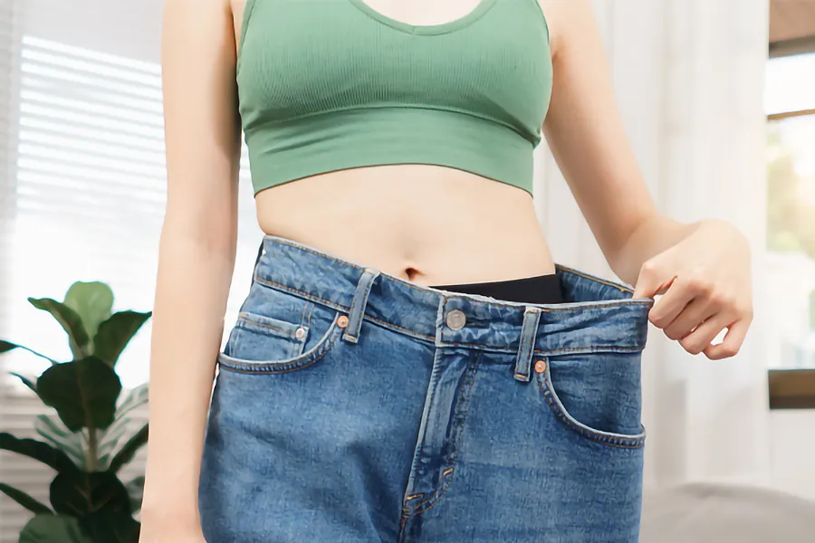 A close-up of a young woman in a green crop top, holding out her oversized blue jeans to show significant weight loss. Zepbound Weight Loss Injections offered by Tanya Zboril, NP of TBT Medical in Edmonton.